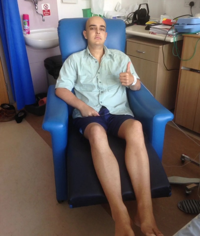 Nick Barnard, bald and wearing a light green shirt and dark blue shorts, gives a thumbs-up from a hospital chair.