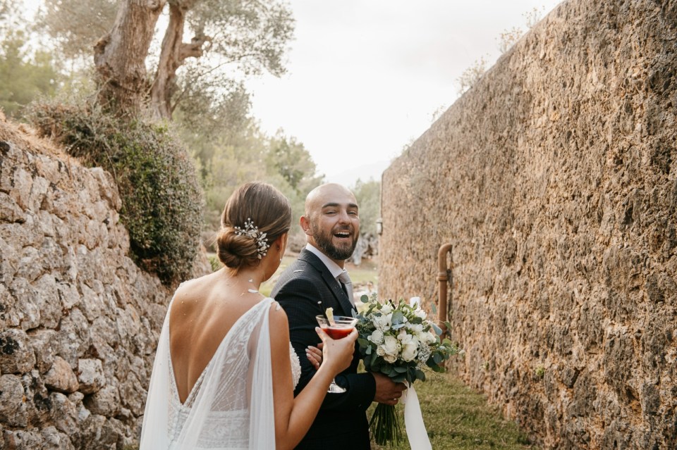 A man identified as Nick Barnard, holding a bouquet and cocktail, laughs while looking back at a woman in a wedding dress.