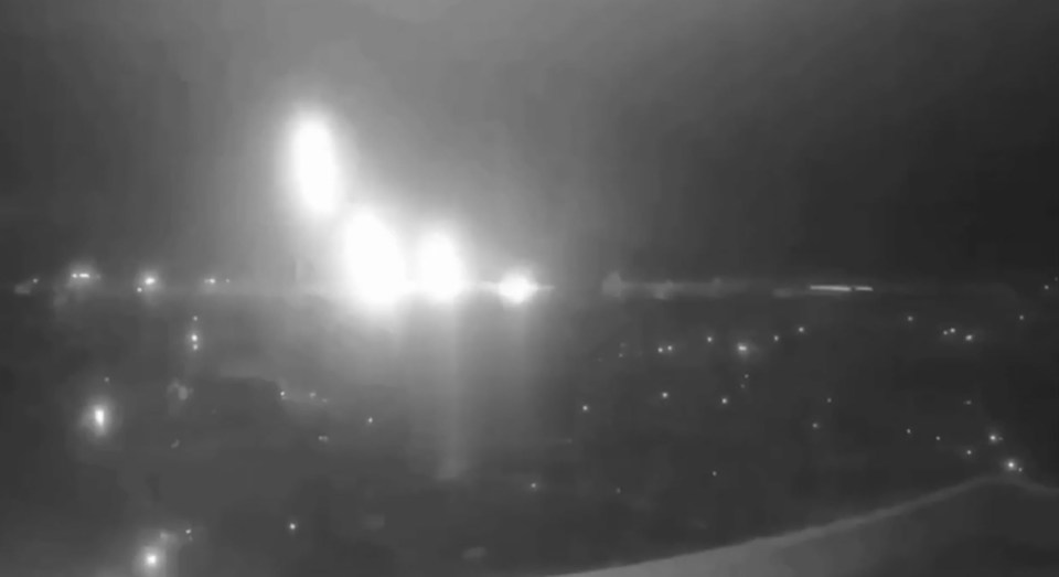 Nighttime camera footage of a large explosion with a bright flash of light over a city.