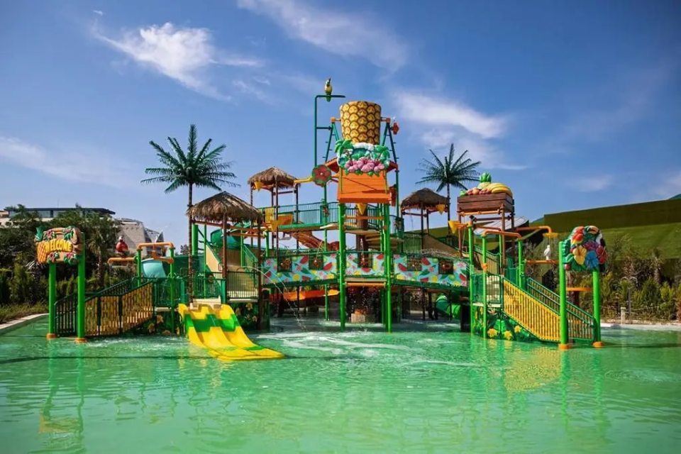 A tropical-themed water park with slides, water features, and palm trees under a blue sky.