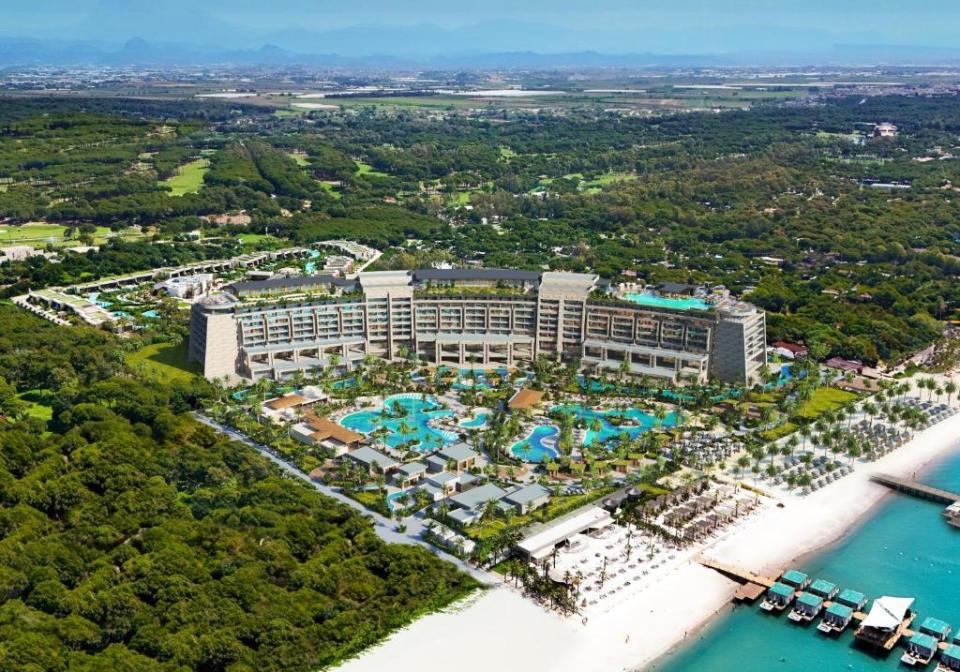 Aerial view of Regnum The Crown, a large resort hotel in Turkey, with pools, a beach, and lush green surroundings.