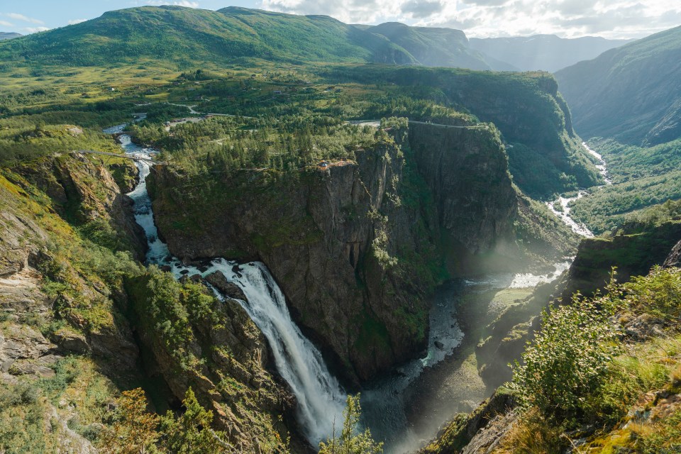 Scenic view of Vøringfossen waterfall in Norway, with cliffs, green trees, and distant mountains.