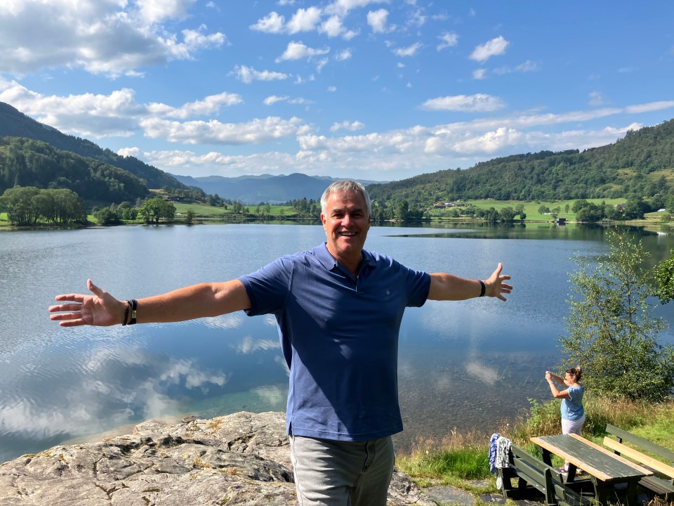 Shaun Custis, a man in a blue polo shirt and grey pants, stands with arms outstretched in front of a lake.