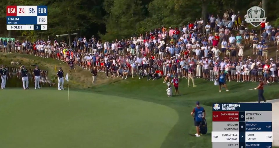 Golf match showing players and caddies on the green with spectators on a hill in the background. Scores of various teams are displayed on a scoreboard.