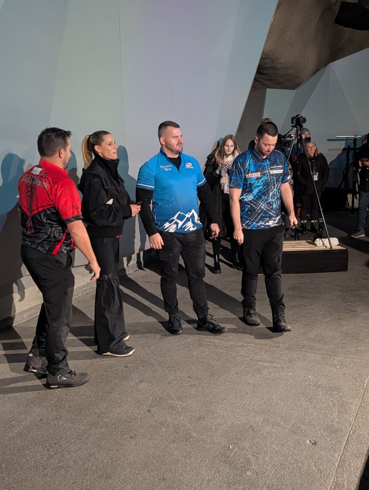 Darts players and a woman standing in a mountain train station with camera equipment in the background.