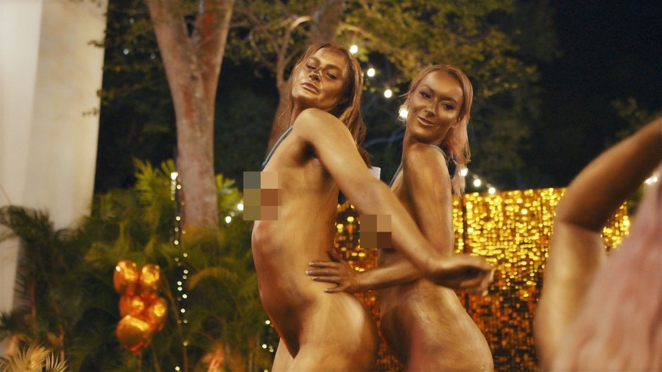 Two women painted gold, posed outdoors at night, with gold balloons and sparkling lights in the background.
