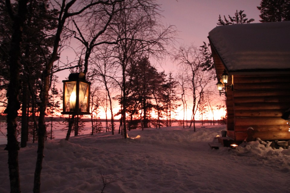 Lanterns light a snowy path leading to a log cabin at sunset in Lapland.