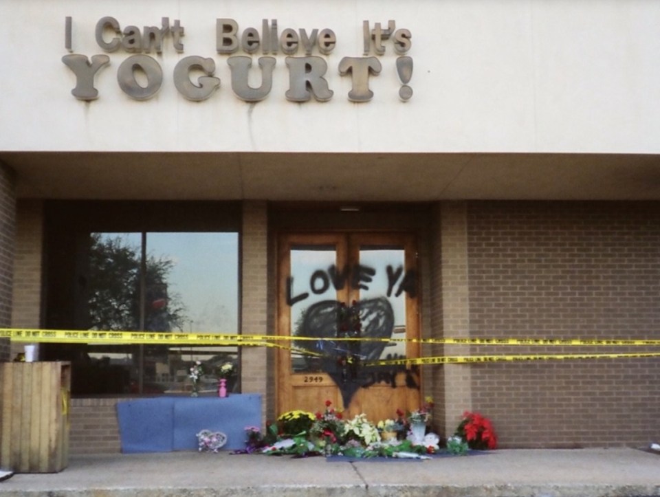 The storefront of an "I Can't Believe It's YOGURT!" shop with police tape, flowers, and "LOVE YA" spray-painted on the door.