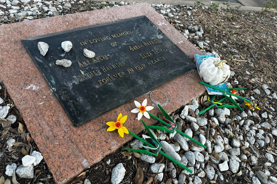 Memorial plaque for Amy Ayers, Sarah Harbison, Jennifer Harbison, and Eliza Thomas, surrounded by tributes including decorative stones, a white pumpkin, and artificial flowers.