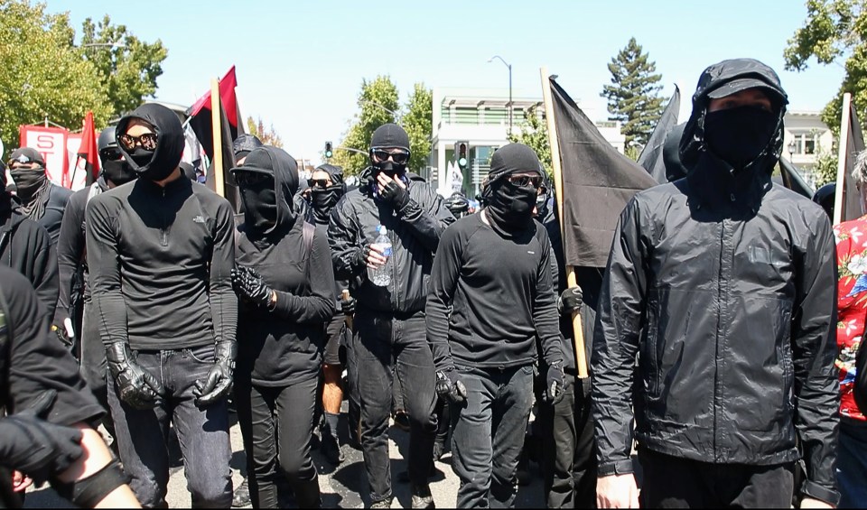 Antifa counter-protesters in black clothing and face coverings march with flags.
