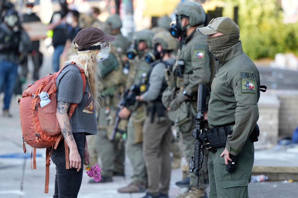 A woman with blonde hair, wearing a cap, goggles, and a respirator mask, confronts a law enforcement officer while holding flowers.