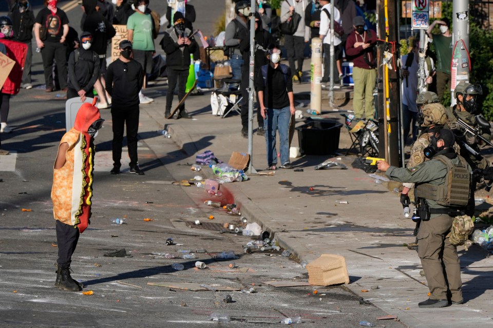 A law enforcement officer in tactical gear points a taser at a person wearing a hot dog costume during a protest.