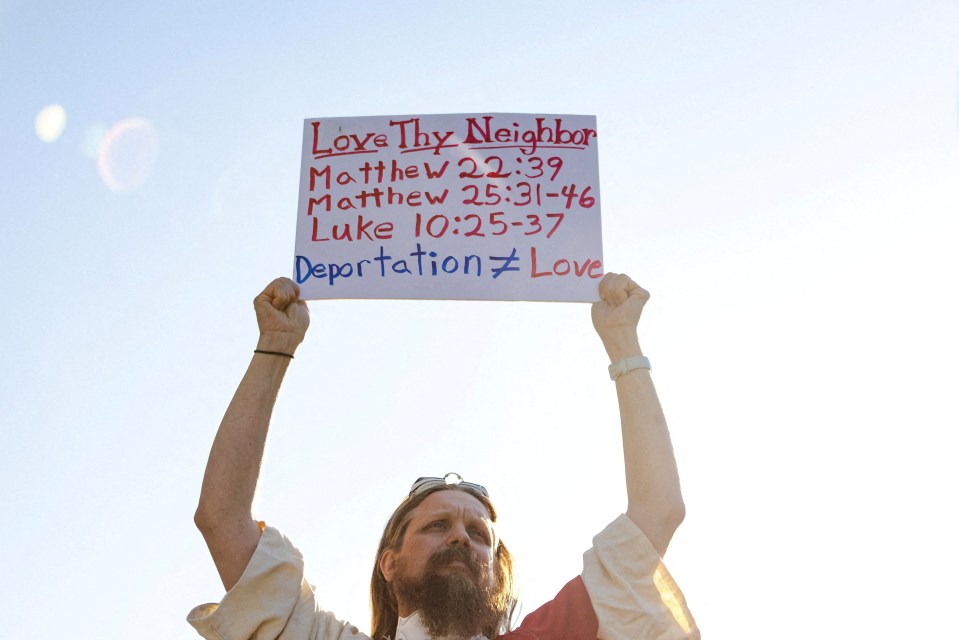 A protester dressed as Jesus holds a sign that reads "Love Thy Neighbor Matthew 22:39 Matthew 25:31-46 Luke 10:25-37 Deportation ≠ Love."