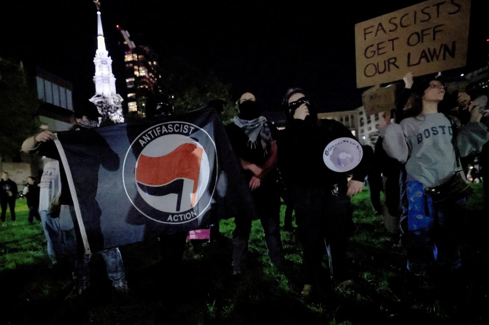 Counter-protesters with an "Antifascist Action" flag and a "Fascists Get Off Our Lawn" sign at night.