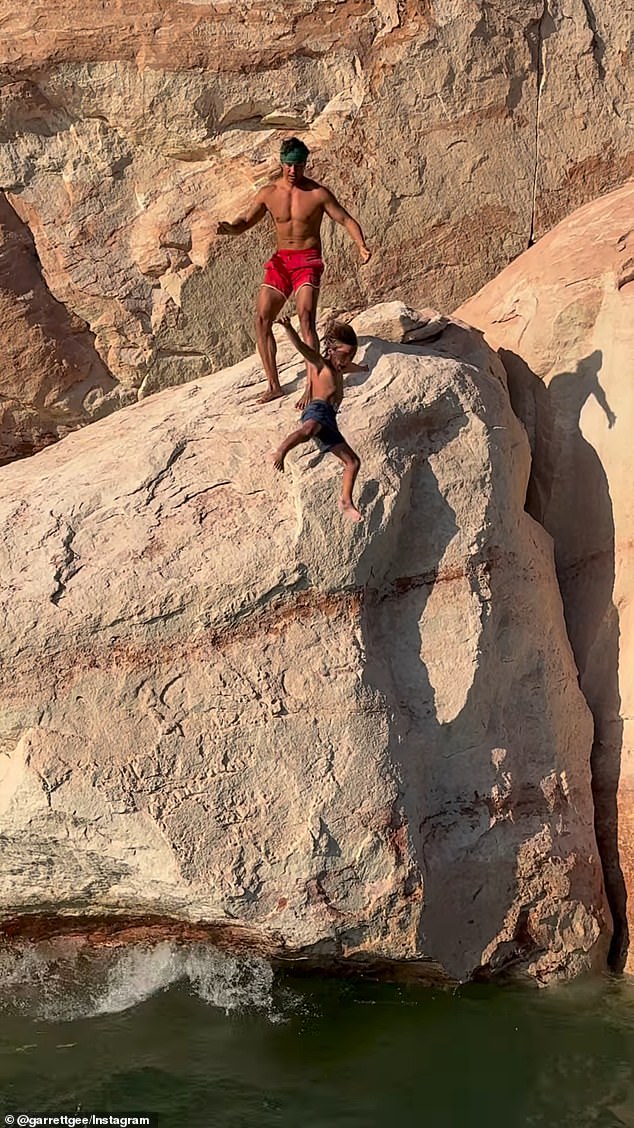 The clip shows Gee standing on a high cliff with his elementary-aged son, Calihan, who appears nervous, shielding his face and turning away from the edge