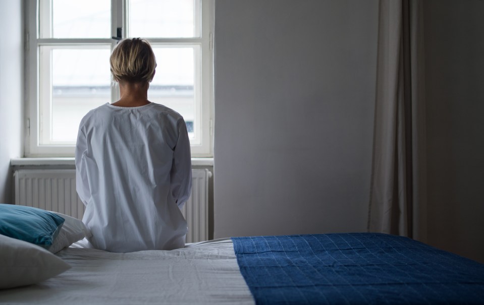 Woman sitting on a bed, facing away from the camera, looking out a window.