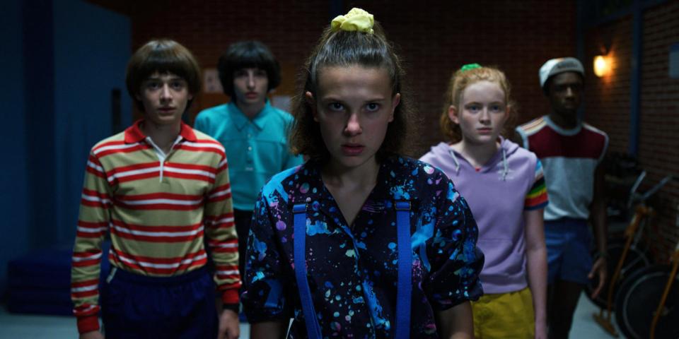 Illustration of the cast of Stranger Things looking concerned, with a haunted house below them and a split red and blue background.