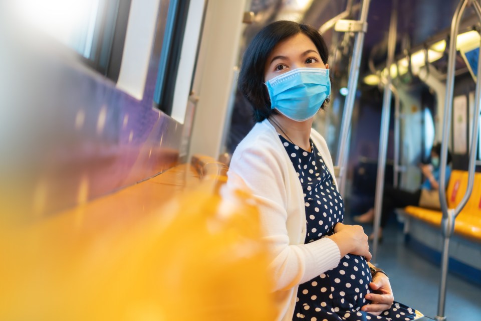 Pregnant Asian woman in a face mask touching her belly on a bus.