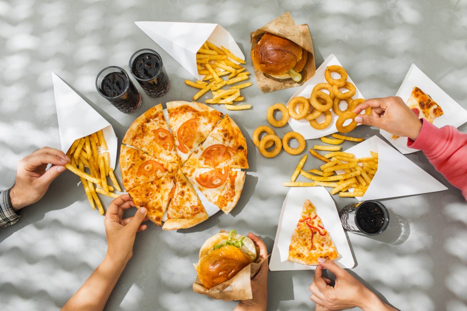 Flat-lay of friends eating burgers, fries, onion rings, pizza, and drinking cola.
