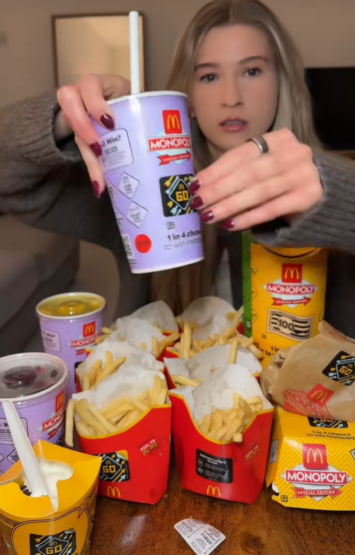 A person holding a McDonald's Monopoly special edition drink cup with other McDonald's items like fries and a dessert.