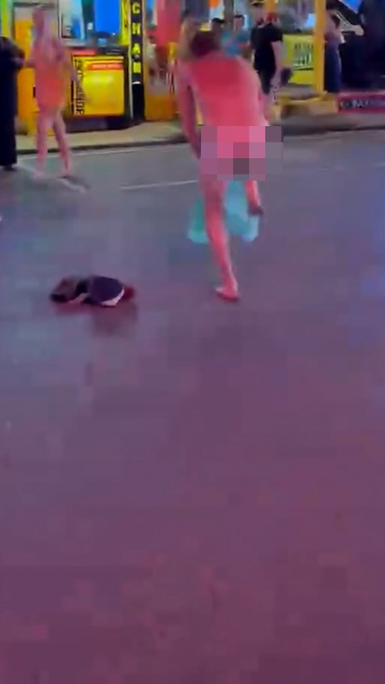 A naked man with pixelated buttocks dances in the middle of a street at night while people watch.