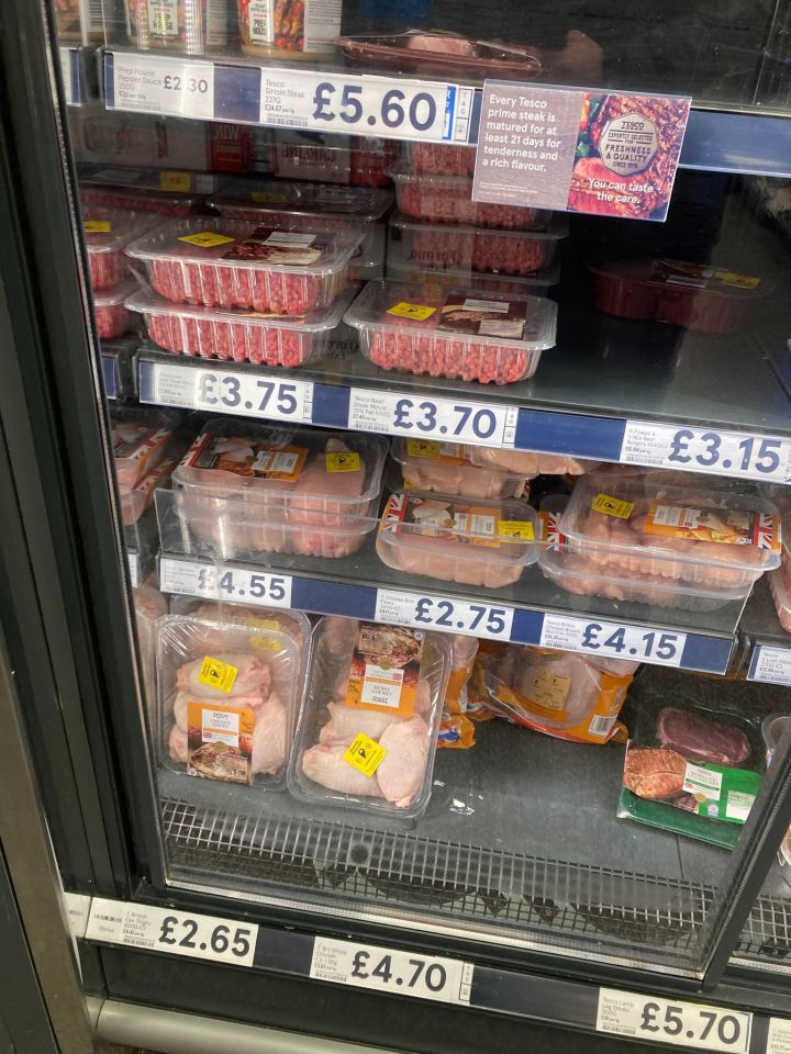 A refrigerated display case in a grocery store filled with various packaged meats, some with security tags attached, and price labels.