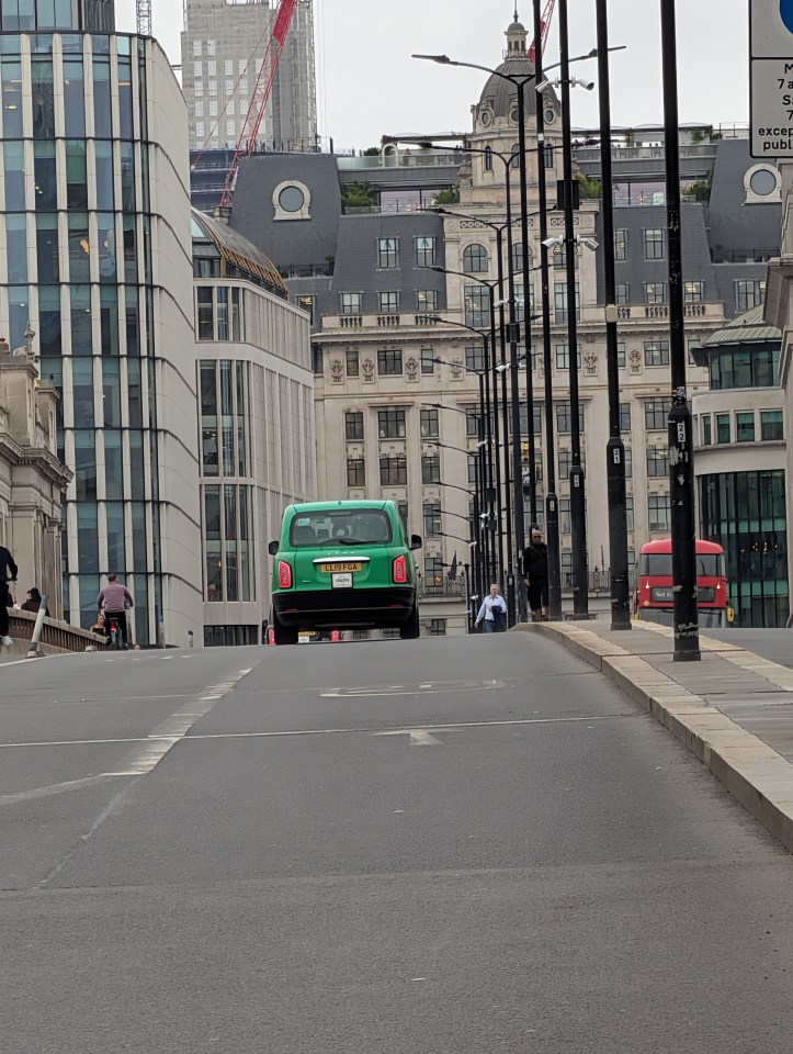 A green London taxi drives away from the camera up a hill, passing by tall modern and older buildings and streetlights.