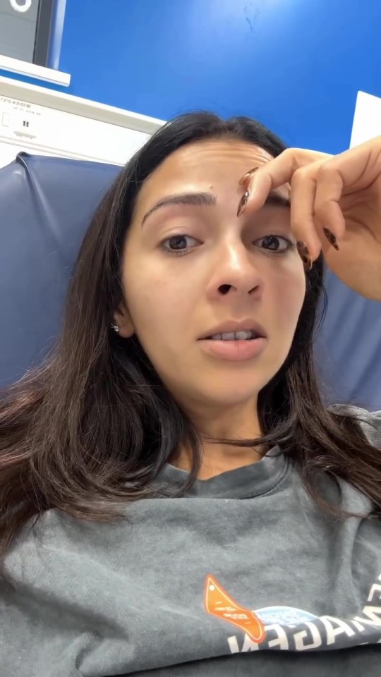 A woman with dark hair, wearing a gray t-shirt, looking concerned and touching her forehead.