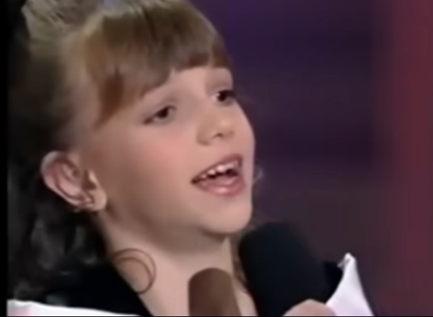 A young female singer with light brown hair and bangs, holding a microphone.
