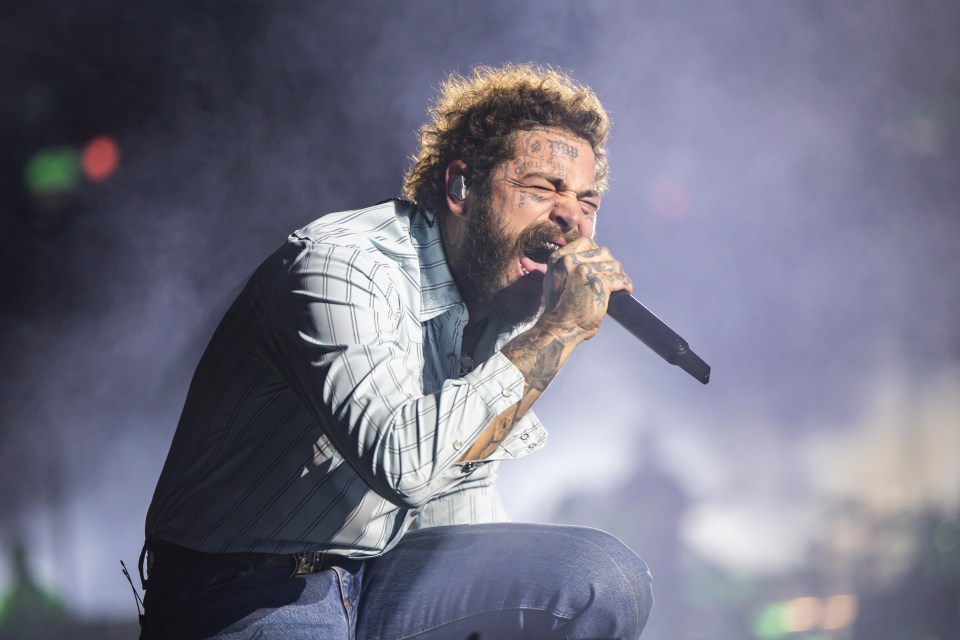 Post Malone performs in concert, wearing a striped shirt and jeans, holding a microphone.