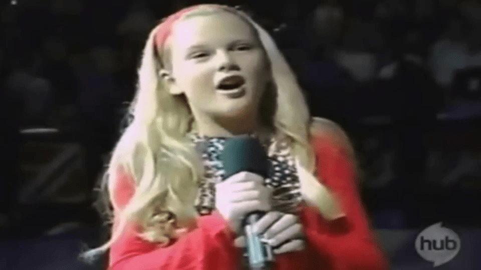 A young Taylor Swift with long blonde hair and a red headband, wearing a red top and patterned dress, singing into a microphone.