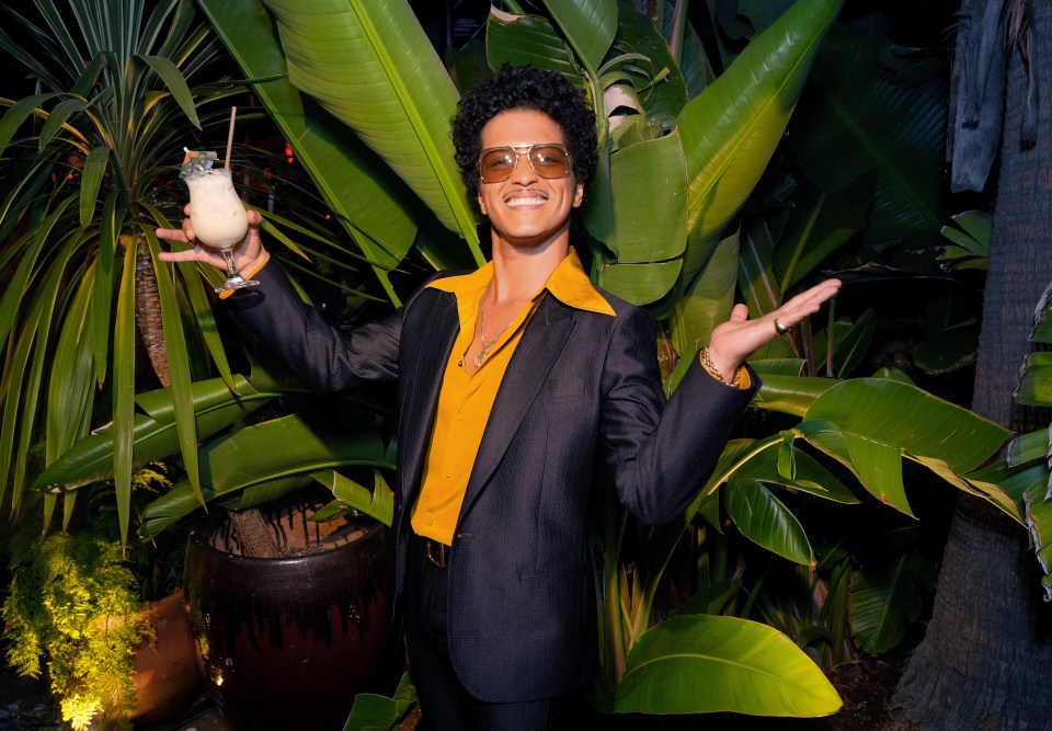 Bruno Mars, co-owner of SelvaRey Rum, wearing sunglasses and a yellow shirt, holding a drink and smiling.
