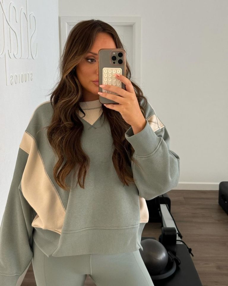 Woman with long brown hair wearing a two-tone sweatshirt and leggings, taking a mirror selfie with a phone.