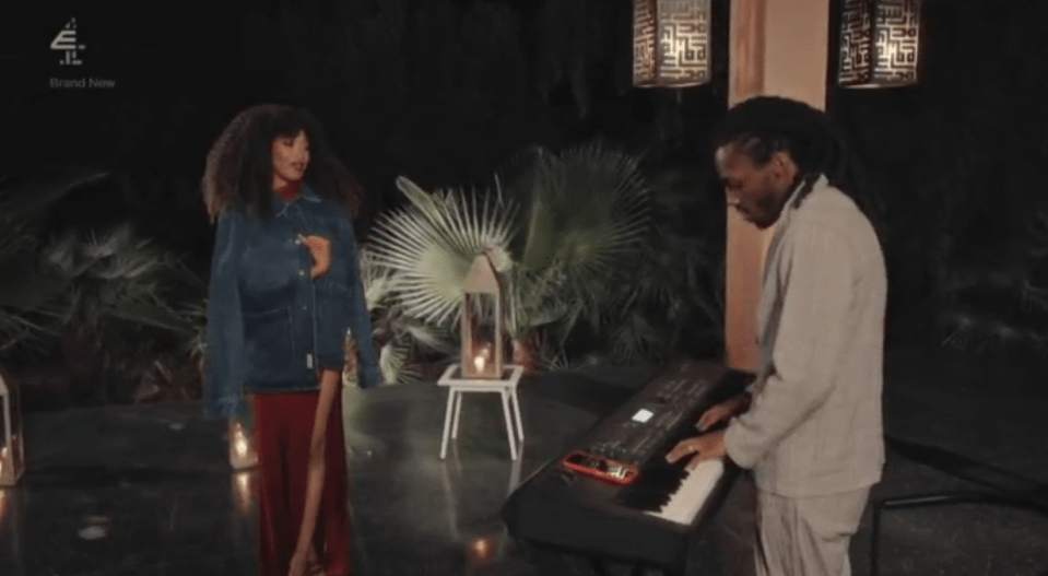 A woman in a blue jacket and red dress stands next to a man playing a keyboard.