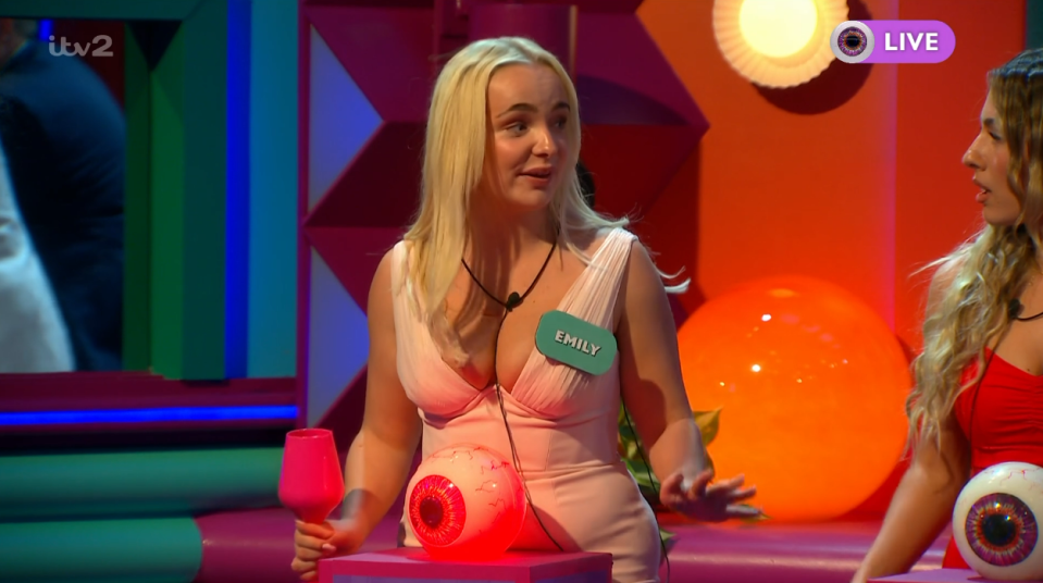 Emily in a pink dress, wearing a name tag, sitting next to a red glowing eyeball in a brightly lit room.