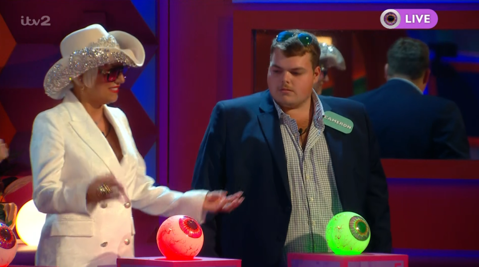 A person in a white suit, sparkly cowboy hat, and sunglasses talks to a man in a blue blazer next to two glowing eyeball orbs.