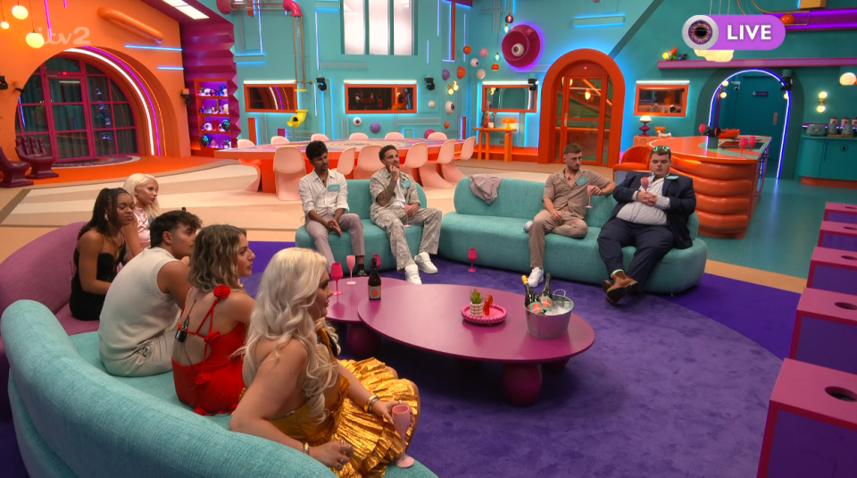 A group of 12 young people, some sitting on a turquoise couch and some on a light purple couch, are gathered in a brightly colored living room.