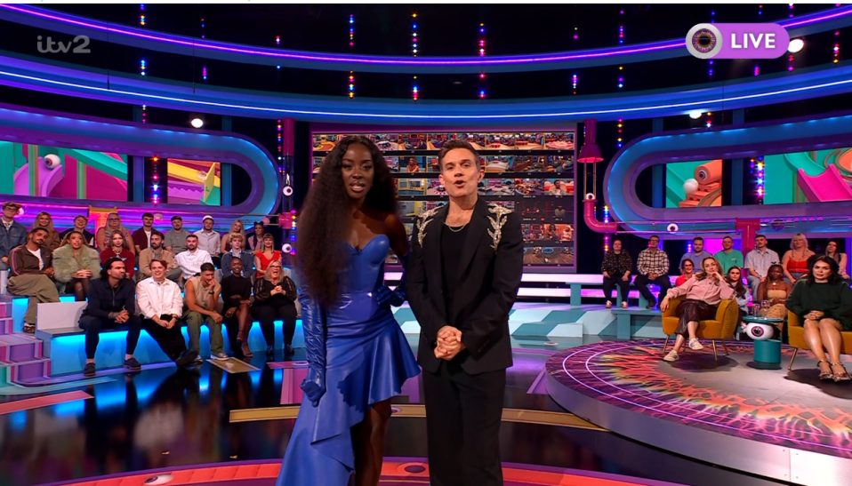 A woman in a blue dress and a man in a black suit hosting a live show with an audience.