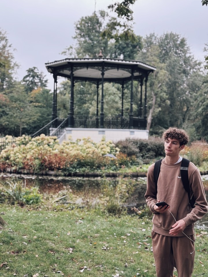 Young man in a brown tracksuit holding a phone with a charging cable, standing in a park with a bandstand in the background.