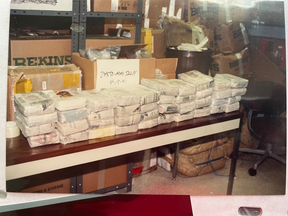 Bundles of drugs wrapped in paper and plastic sitting on a long table in a room full of boxes.