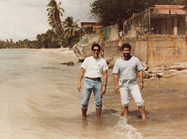 Two men standing in the ocean on a beach with palm trees and buildings in the background.
