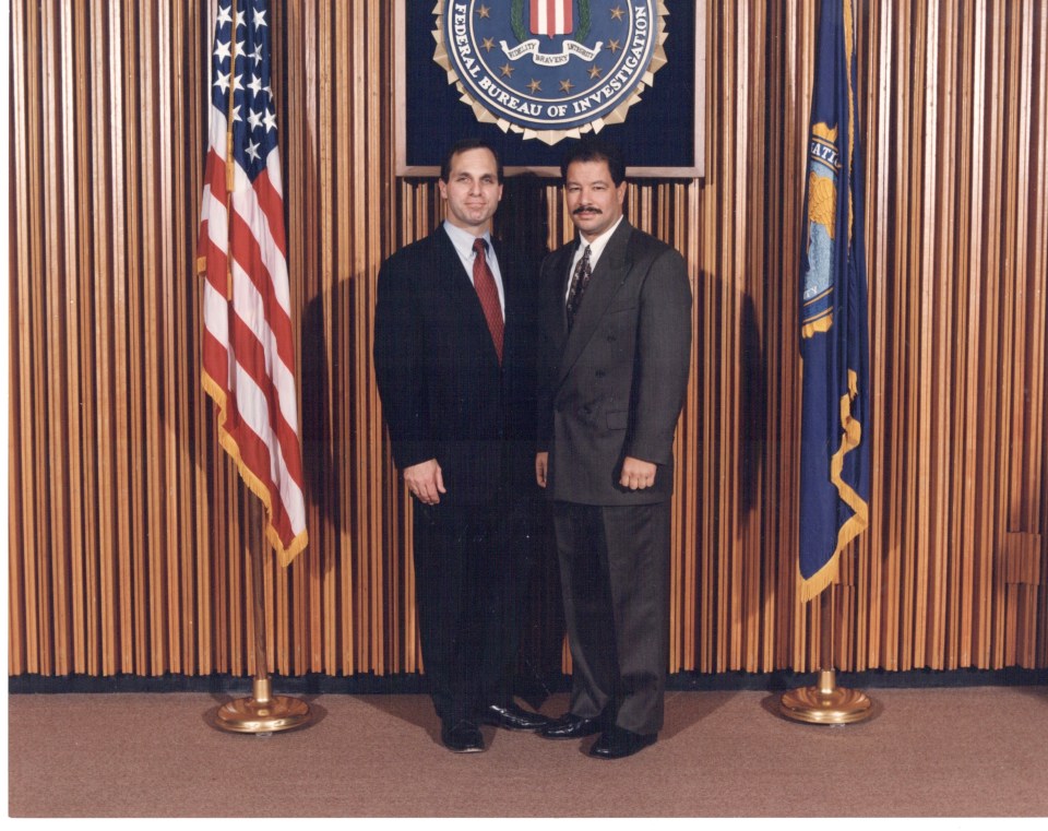 Martin Suarez and another man in suits standing in front of an FBI seal and two flags.