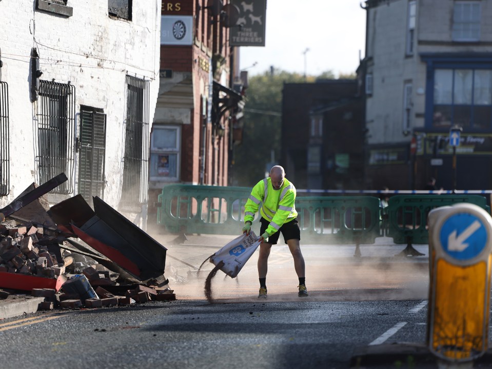 A man in a neon safety vest pours sand on the street after a car crashed into a building.