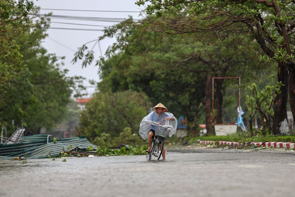 A woman in a conical hat and clear poncho rides a bicycle through a flooded street during Typhoon Bualoi in Vietnam.