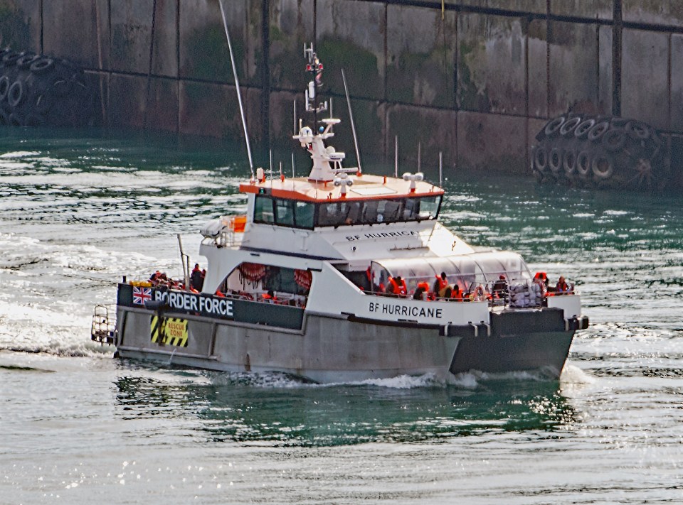 A Border Force vessel, "BF Hurricane," with rescued migrants in orange life jackets, moving through water at Dover Docks.