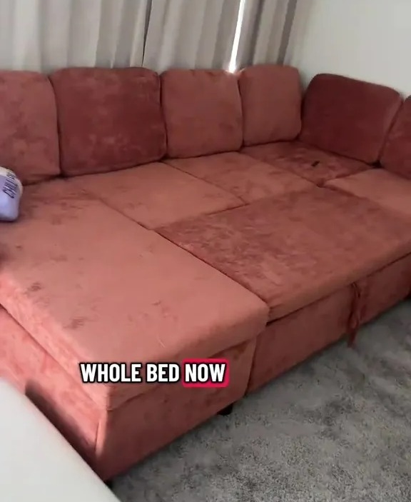 A pink sectional sofa unfolded into a bed with hidden storage, in front of a window with white curtains.