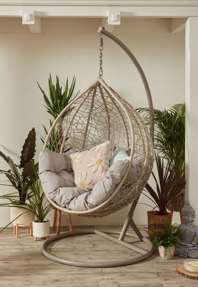 Woven egg chair with gray cushions and a pink daisy pillow, surrounded by potted plants.