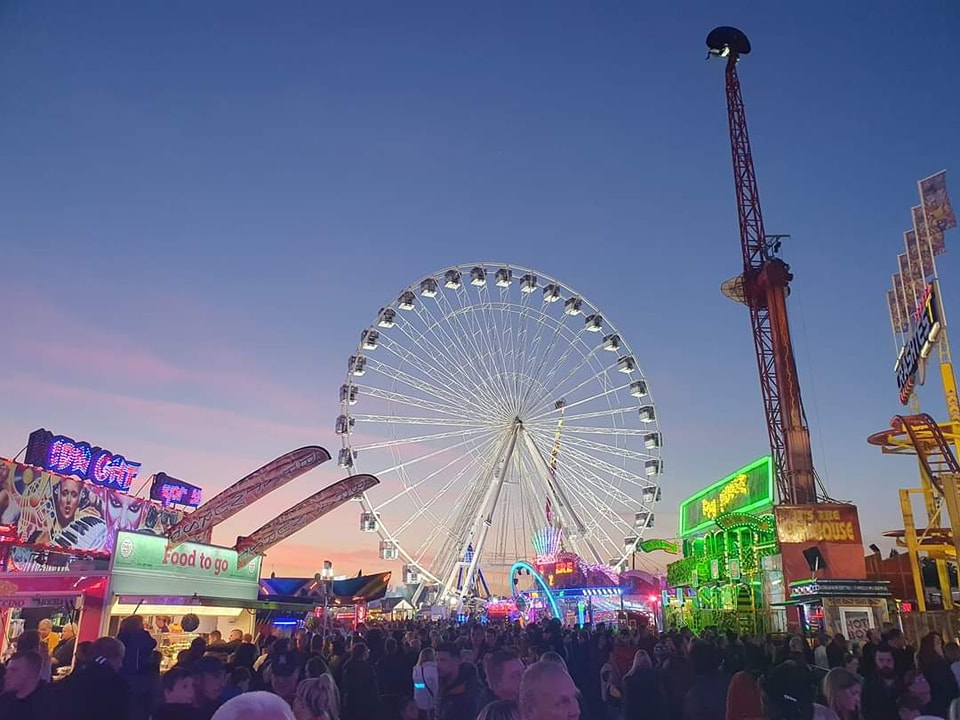 A crowded Hull Fair at dusk with a large Ferris wheel and other illuminated rides.