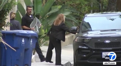 People near a black SUV, two blue trash cans, and green foliage.