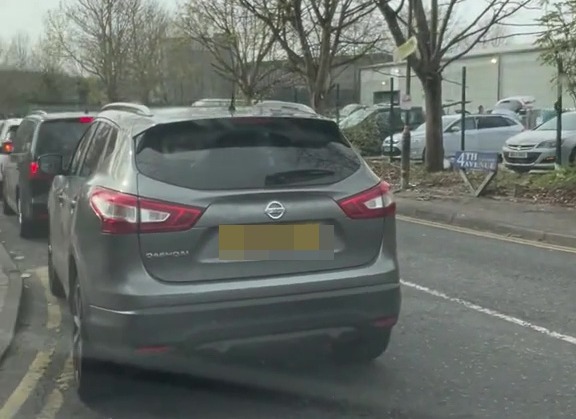 Cars lined up on a street with bare trees overhead, indicating a traffic queue.